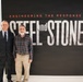 Two men are standing in front of a large exhibition display titled 'Engineering the Response to 9/11: Steel and Stone,' which appears to be a museum or commemorative exhibit about the U.S. Army Corps of Engineers' role in the 9/11 recovery efforts. One man is dressed in a formal dark suit with a tie, while the other wears a casual plaid shirt and khaki pants. The exhibit panel includes descriptive text about USACE's involvement at the World Trade Center and Pentagon sites.