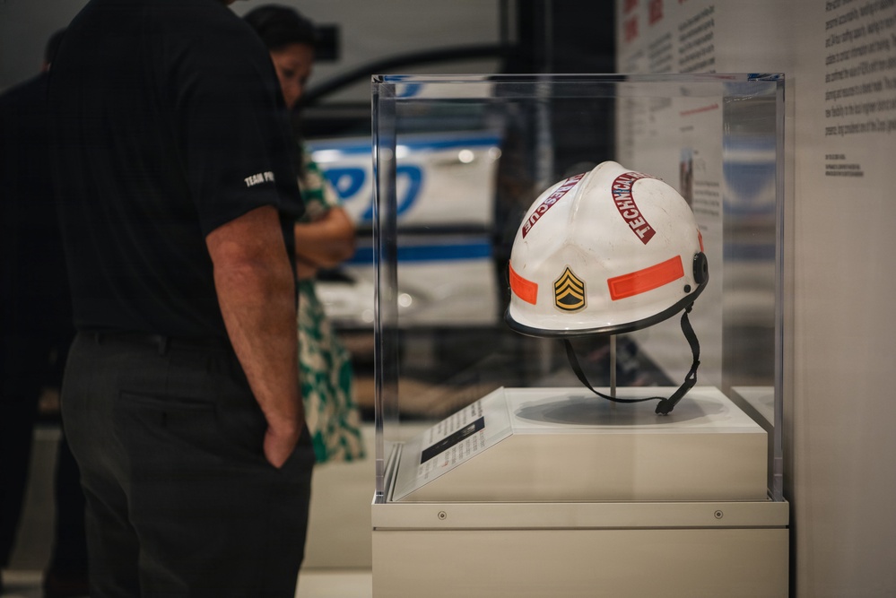 A white helmet is displayed in a clear acrylic museum case on a pedestal. The helmet features orange reflective strips, a military sergeant chevron patch, and text reading 'TECHNICAL' on the front. Two museum visitors are visible in the blurred background examining another exhibit.
