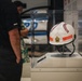A white helmet is displayed in a clear acrylic museum case on a pedestal. The helmet features orange reflective strips, a military sergeant chevron patch, and text reading 'TECHNICAL' on the front. Two museum visitors are visible in the blurred background examining another exhibit.