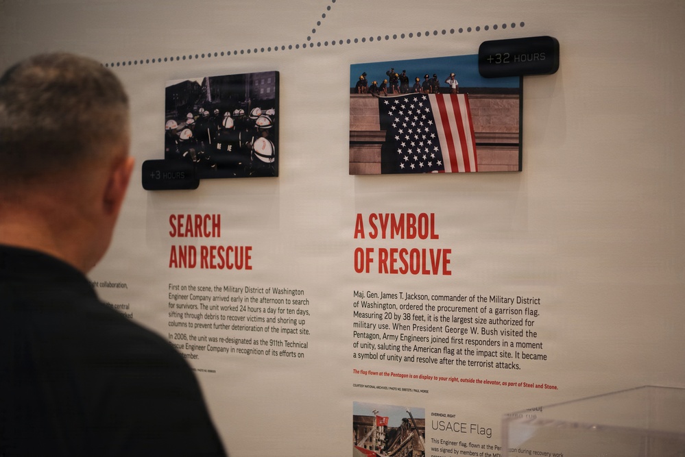 A museum exhibit display showing two panels related to the September 11, 2001 Pentagon attack. The panels are titled 'Search and Rescue' (+3 Hours) and 'A Symbol of Resolve' (+32 Hours), featuring historical photographs and descriptive text. A man with gray hair, viewed from behind, is reading the exhibit wall.