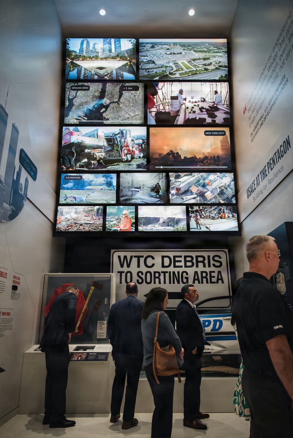 The image shows visitors viewing a museum exhibit related to the September 11, 2001 attacks, featuring a large multi-screen video wall displaying archival footage and photographs of Ground Zero, the Pentagon, and New York City. A prominent 'WTC Debris to Sorting Area' sign is displayed alongside a damaged NYPD vehicle. The exhibit appears to be part of the 9/11 Memorial Museum or a related USACE (U.S. Army Corps of Engineers) traveling exhibition titled 'USACE at the Pentagon: Building for Resilience.'