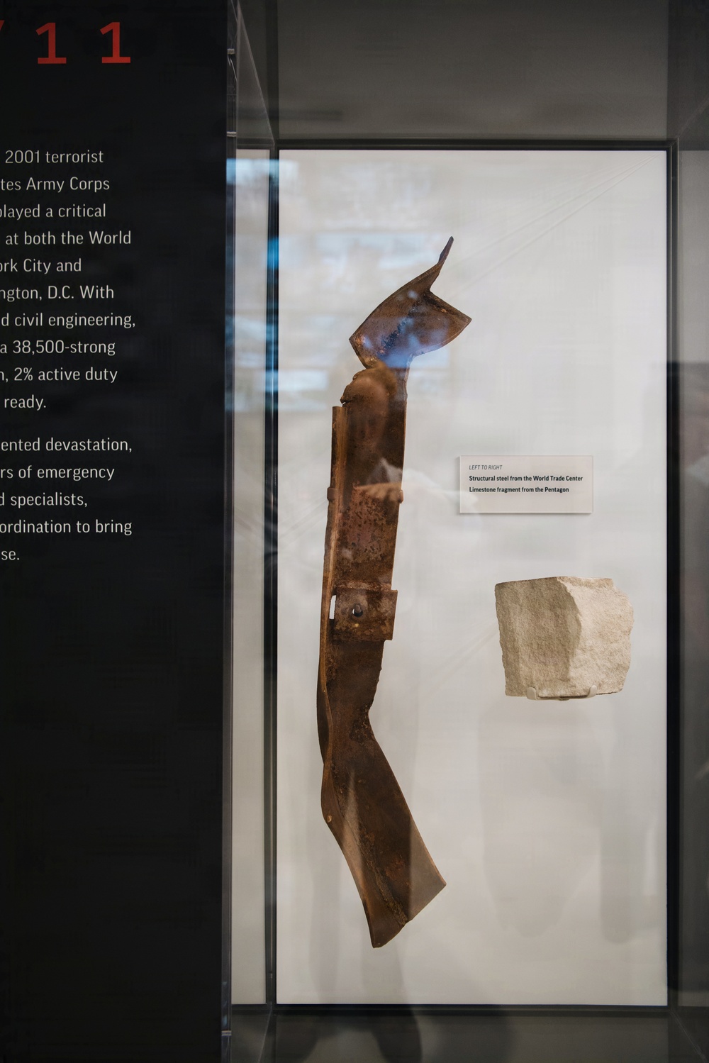 A museum display case contains two artifacts related to the September 11, 2001 terrorist attacks: a twisted, heavily corroded structural steel beam from the World Trade Center and a limestone fragment from the Pentagon. The exhibit label reads 'LEFT TO RIGHT: Structural steel from the World Trade Center / Limestone fragment from the Pentagon.' Partial wall text on the left references the 2001 terrorist attacks and the United States Army Corps of Engineers' response role.
