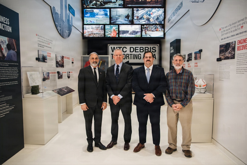Four men are posing for a formal group photograph inside what appears to be a 9/11 memorial or museum exhibit. The backdrop includes a 'WTC Debris Reporting Area' sign, multiple display screens showing images of destruction and rescue operations, and informational panels about the September 11 attacks. Three of the men are wearing dark business suits while one is dressed in casual plaid attire.