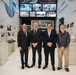 Four men are posing for a formal group photograph inside what appears to be a 9/11 memorial or museum exhibit. The backdrop includes a 'WTC Debris Reporting Area' sign, multiple display screens showing images of destruction and rescue operations, and informational panels about the September 11 attacks. Three of the men are wearing dark business suits while one is dressed in casual plaid attire.