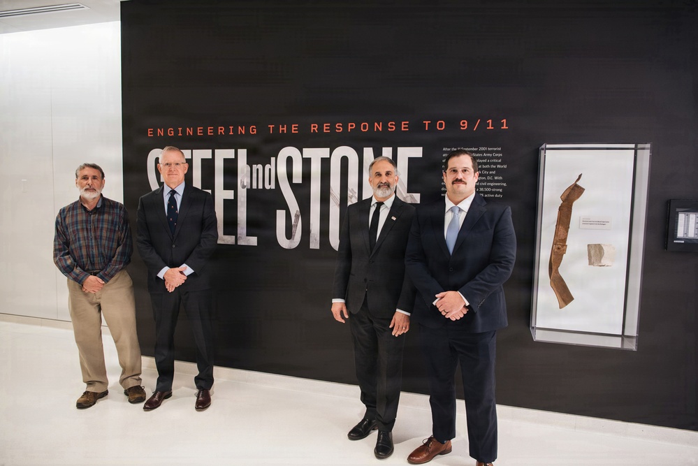 Four men are posing in front of a large exhibition display titled 'Steel and Stone: Engineering the Response to 9/11.' The setting appears to be a museum or exhibit hall with a modern, clean interior. Three of the men are wearing formal suits while one is dressed in casual attire.