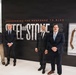 Four men are posing in front of a large exhibition display titled 'Steel and Stone: Engineering the Response to 9/11.' The setting appears to be a museum or exhibit hall with a modern, clean interior. Three of the men are wearing formal suits while one is dressed in casual attire.