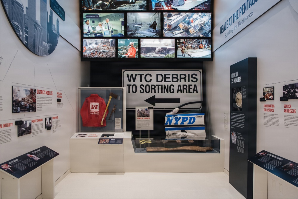 This image shows a museum exhibition dedicated to the September 11, 2001 attacks and recovery efforts, specifically focused on USACE (U.S. Army Corps of Engineers) operations at the Pentagon and World Trade Center. The display features authentic artifacts, signage, and multimedia screens documenting rescue, recovery, and forensic operations. The exhibit is professionally curated with labeled display cases, informational panels, and archival photographs.