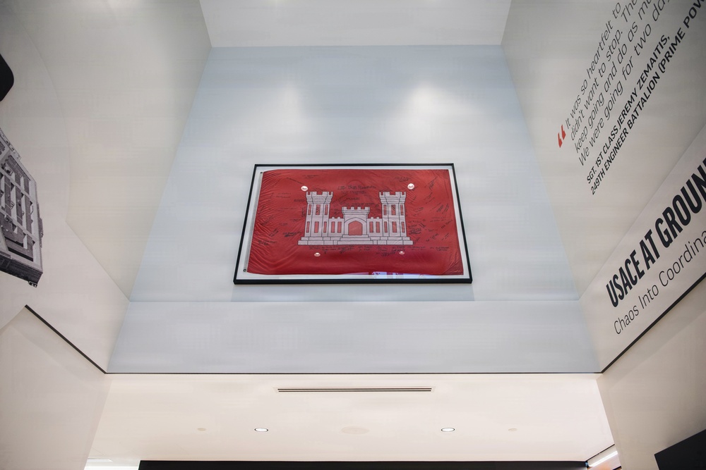The image shows a museum or exhibition space featuring a framed red flag with a white castle emblem — the insignia of the U.S. Army Corps of Engineers (USACE) — mounted high on a light-colored wall. The flag appears to be signed by multiple individuals and is displayed as an artifact. Surrounding wall text references 'USACE AT GROUND ZERO' and includes a quote attributed to SGT 1ST CLASS JEREMY ZEMAITIS, 249TH ENGINEER BATTALION.