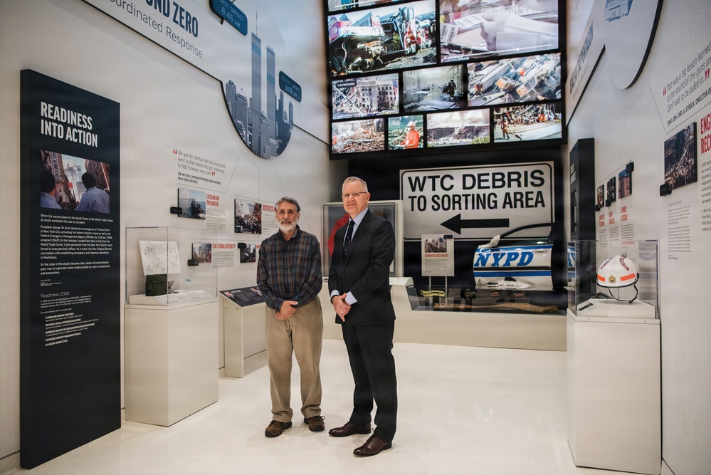 Two men stand inside what appears to be a 9/11 memorial museum exhibit. The exhibit features artifacts and signage related to the World Trade Center attacks and recovery efforts, including an NYPD vehicle and a 'WTC Debris to Sorting Area' directional sign. The display includes multiple video screens showing archival footage and a panel titled 'Readiness Into Action.'
