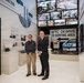 Two men stand inside what appears to be a 9/11 memorial museum exhibit. The exhibit features artifacts and signage related to the World Trade Center attacks and recovery efforts, including an NYPD vehicle and a 'WTC Debris to Sorting Area' directional sign. The display includes multiple video screens showing archival footage and a panel titled 'Readiness Into Action.'