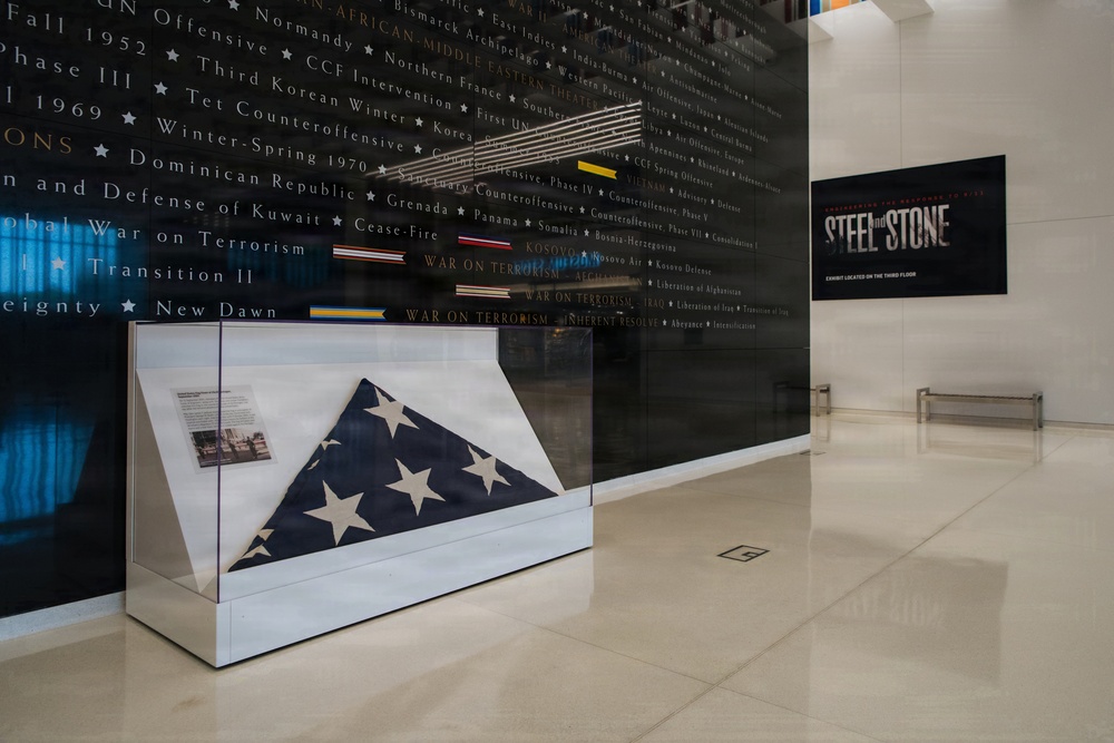 A museum exhibit hall featuring a glass display case containing a folded American flag (tri-corner military funeral fold). Behind the display case is a large dark wall engraved with military campaigns and conflicts spanning from the Korean War era through modern operations. A sign on the right wall advertises an exhibit called 'Steel and Stone' located on the third floor.