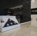 A museum exhibit hall featuring a glass display case containing a folded American flag (tri-corner military funeral fold). Behind the display case is a large dark wall engraved with military campaigns and conflicts spanning from the Korean War era through modern operations. A sign on the right wall advertises an exhibit called 'Steel and Stone' located on the third floor.