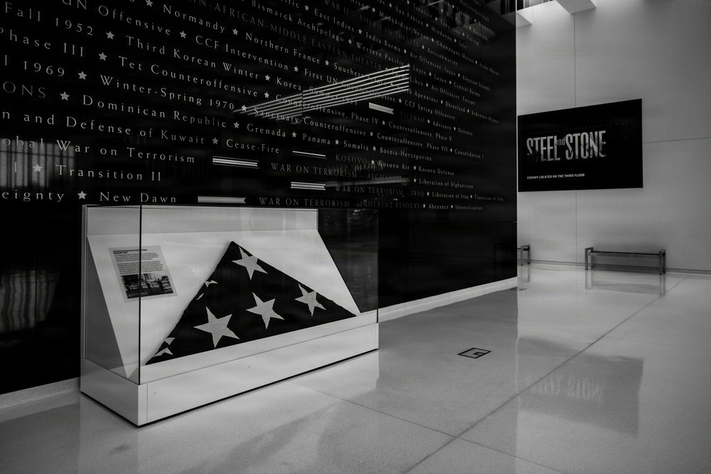 A black-and-white photograph of a military museum interior featuring a folded American flag displayed in a glass case. Behind the display case is a large dark wall inscribed with military campaigns and conflicts spanning from World War II through modern operations. A sign on the right wall advertises an exhibit called 'Steel and Stone' located on the third floor.
