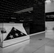 A black-and-white photograph of a military museum interior featuring a folded American flag displayed in a glass case. Behind the display case is a large dark wall inscribed with military campaigns and conflicts spanning from World War II through modern operations. A sign on the right wall advertises an exhibit called 'Steel and Stone' located on the third floor.