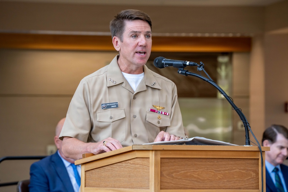 A military officer in U.S. Navy Service Uniform (tan/khaki) is speaking at a wooden podium with a microphone. His name tag reads 'John Dougherty III' with the rank of Commander. He appears to be delivering a speech or remarks at a formal event, with civilians visible in the background.