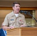 A military officer in U.S. Navy Service Uniform (tan/khaki) is speaking at a wooden podium with a microphone. His name tag reads 'John Dougherty III' with the rank of Commander. He appears to be delivering a speech or remarks at a formal event, with civilians visible in the background.