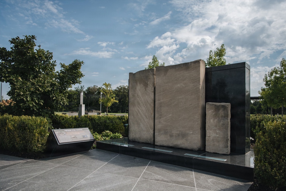 The image shows an outdoor memorial installation featuring large stone slabs mounted against a dark reflective black panel, set within a landscaped garden environment. A descriptive placard on the left reads 'Pentagon Stone Memorial,' indicating this is a commemorative site. The memorial is surrounded by manicured hedges, trees, and a polished stone plaza under a partly cloudy sky.