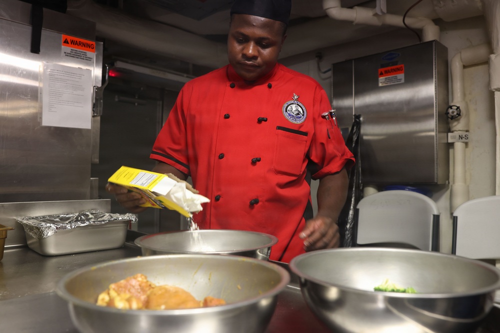 A chef wearing a red uniform with an embroidered badge and black cap is preparing food in what appears to be a professional or institutional kitchen. He is pouring contents from a yellow box into a stainless steel mixing bowl, with multiple bowls containing various food items visible in the foreground. The setting appears to be a military or naval galley based on the industrial equipment and warning labels visible in the background.