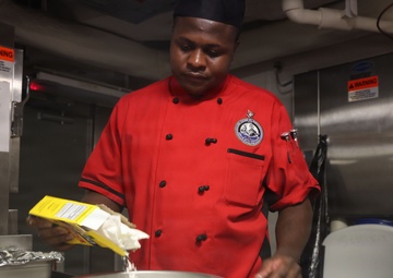 A chef wearing a red uniform with an embroidered badge and black cap is preparing food in what appears to be a professional or institutional kitchen. He is pouring contents from a yellow box into a stainless steel mixing bowl, with multiple bowls containing various food items visible in the foreground. The setting appears to be a military or naval galley based on the industrial equipment and warning labels visible in the background.