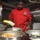 A chef wearing a red uniform with an embroidered badge and black cap is preparing food in what appears to be a professional or institutional kitchen. He is pouring contents from a yellow box into a stainless steel mixing bowl, with multiple bowls containing various food items visible in the foreground. The setting appears to be a military or naval galley based on the industrial equipment and warning labels visible in the background.