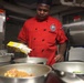 A chef wearing a red uniform with an embroidered badge and black cap is preparing food in what appears to be a professional or institutional kitchen. He is pouring contents from a yellow box into a stainless steel mixing bowl, with multiple bowls containing various food items visible in the foreground. The setting appears to be a military or naval galley based on the industrial equipment and warning labels visible in the background.