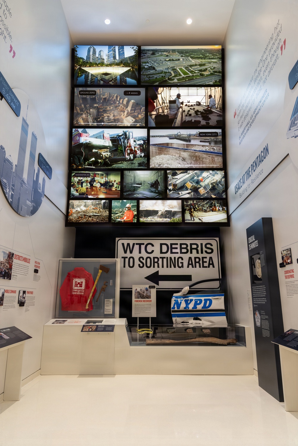 This image shows a museum exhibit dedicated to the September 11, 2001 attacks and their aftermath, focusing on recovery and response efforts. The display features a prominent 'WTC DEBRIS TO SORTING AREA' directional sign, physical artifacts in glass cases, and a large video wall showing multiple photographs of Ground Zero, the Pentagon, and recovery operations. The exhibit appears to be part of a U.S. Army Corps of Engineers (USACE) installation titled 'USACE at the Pentagon: Building for Resilience.'