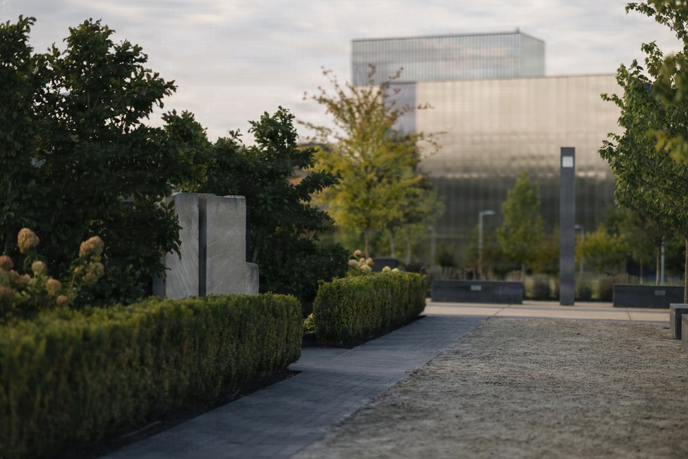 A formal landscape garden featuring neatly trimmed hedgerows, ornamental plantings, and a stone or concrete sculptural element in the foreground. A modern glass-and-steel building is visible in the soft-focus background, suggesting an institutional or corporate campus setting. The image is taken during golden hour or overcast daylight, giving the scene a calm, refined atmosphere.