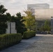 A formal landscape garden featuring neatly trimmed hedgerows, ornamental plantings, and a stone or concrete sculptural element in the foreground. A modern glass-and-steel building is visible in the soft-focus background, suggesting an institutional or corporate campus setting. The image is taken during golden hour or overcast daylight, giving the scene a calm, refined atmosphere.