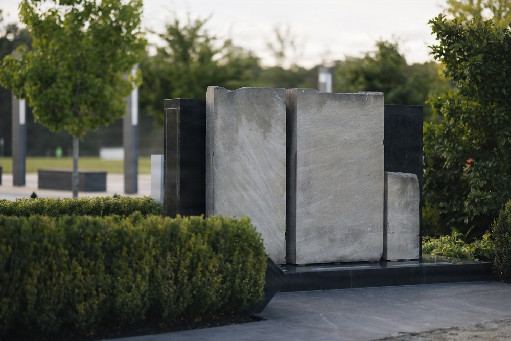 The image shows a modern outdoor memorial or sculptural installation featuring large upright stone slabs mounted on a dark granite base. The sculpture consists of light gray stone panels with natural veining, set against a dark polished stone backdrop. The setting is a well-maintained park or memorial garden with trimmed hedges, trees, and paved pathways.