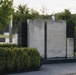 The image shows a modern outdoor memorial or sculptural installation featuring large upright stone slabs mounted on a dark granite base. The sculpture consists of light gray stone panels with natural veining, set against a dark polished stone backdrop. The setting is a well-maintained park or memorial garden with trimmed hedges, trees, and paved pathways.