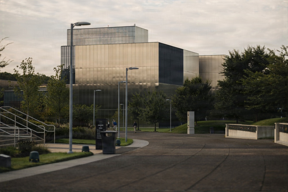 The image shows a large modern institutional building with a distinctive metallic or glass panel facade, surrounded by landscaped grounds with trees and walkways. The architecture features geometric, box-like forms with reflective cladding. The scene appears to be a campus or government/research facility photographed during overcast daylight conditions.