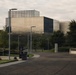 The image shows a large modern institutional building with a distinctive metallic or glass panel facade, surrounded by landscaped grounds with trees and walkways. The architecture features geometric, box-like forms with reflective cladding. The scene appears to be a campus or government/research facility photographed during overcast daylight conditions.