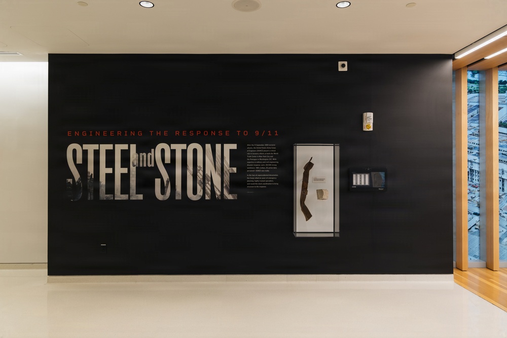 A museum or exhibition installation featuring a large black wall display titled 'Steel and Stone: Engineering the Response to 9/11.' The exhibit includes large typographic wall graphics, a glass display case containing artifacts, and an informational text panel. The installation appears to be in a modern, well-lit gallery space with wood-framed windows visible on the right.