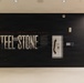 A museum or exhibition installation featuring a large black wall display titled 'Steel and Stone: Engineering the Response to 9/11.' The exhibit includes large typographic wall graphics, a glass display case containing artifacts, and an informational text panel. The installation appears to be in a modern, well-lit gallery space with wood-framed windows visible on the right.