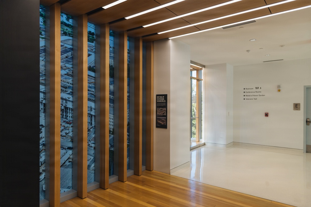 This image shows a modern interior corridor or lobby space featuring warm wood architectural elements and vertical slat panels with large photographic murals depicting an aerial view of what appears to be the Pentagon in Washington, D.C. The space includes directional signage pointing to restrooms, conference rooms, Medal of Honor Garden, and Veterans' Hall, suggesting this is a military or government museum/memorial facility. The design combines natural wood finishes, polished floors, and recessed linear lighting for a contemporary institutional aesthetic.