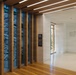 This image shows a modern interior corridor or lobby space featuring warm wood architectural elements and vertical slat panels with large photographic murals depicting an aerial view of what appears to be the Pentagon in Washington, D.C. The space includes directional signage pointing to restrooms, conference rooms, Medal of Honor Garden, and Veterans' Hall, suggesting this is a military or government museum/memorial facility. The design combines natural wood finishes, polished floors, and recessed linear lighting for a contemporary institutional aesthetic.
