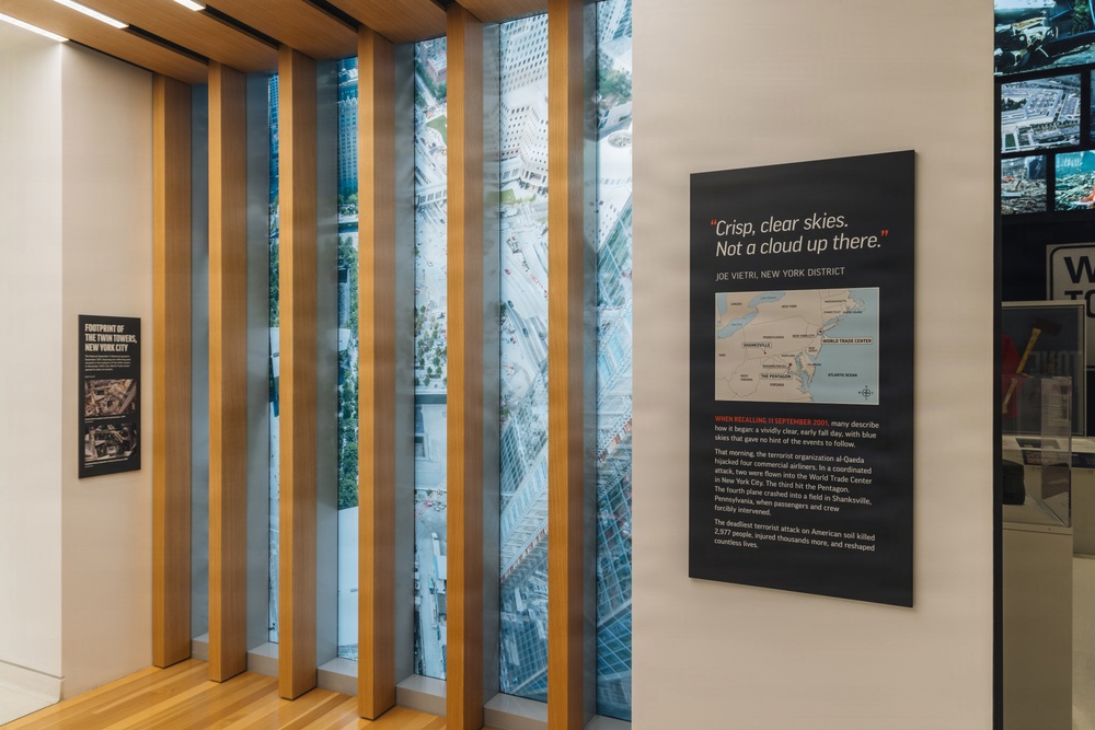 This image shows an interior exhibit space, likely from the 9/11 Memorial Museum in New York City. The display features interpretive panels, a large aerial photograph of lower Manhattan visible through vertical wooden slats, and informational text about the September 11, 2001 terrorist attacks. The exhibit includes a prominent quote from Joe Vietri, New York District, reading 'Crisp, clear skies. Not a cloud up there.'