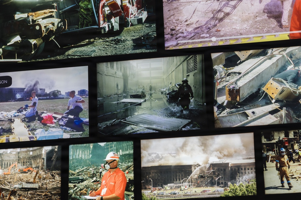A large display wall featuring multiple archival photographs documenting the September 11, 2001 attacks and their aftermath, including scenes from the Pentagon and World Trade Center sites. The photos show emergency responders, rescue operations, structural damage, and debris removal efforts. A museum visitor or worker in an orange shirt and white hard hat with a respirator mask is visible in the foreground, suggesting this is a memorial or museum exhibit.