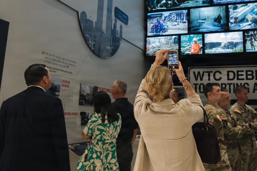 Visitors are touring what appears to be the 9/11 Memorial Museum, as evidenced by visible signage reading 'WTC DEB[RIS]' and display screens showing images of The Pentagon and World Trade Center destruction. A woman in a beige blazer is photographing the video wall with her smartphone, while military personnel in camouflage uniforms and civilians view the exhibits. The museum displays include informational panels, archival photographs, and multiple video screens showing rescue and recovery operations.