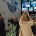 Visitors are touring what appears to be the 9/11 Memorial Museum, as evidenced by visible signage reading 'WTC DEB[RIS]' and display screens showing images of The Pentagon and World Trade Center destruction. A woman in a beige blazer is photographing the video wall with her smartphone, while military personnel in camouflage uniforms and civilians view the exhibits. The museum displays include informational panels, archival photographs, and multiple video screens showing rescue and recovery operations.