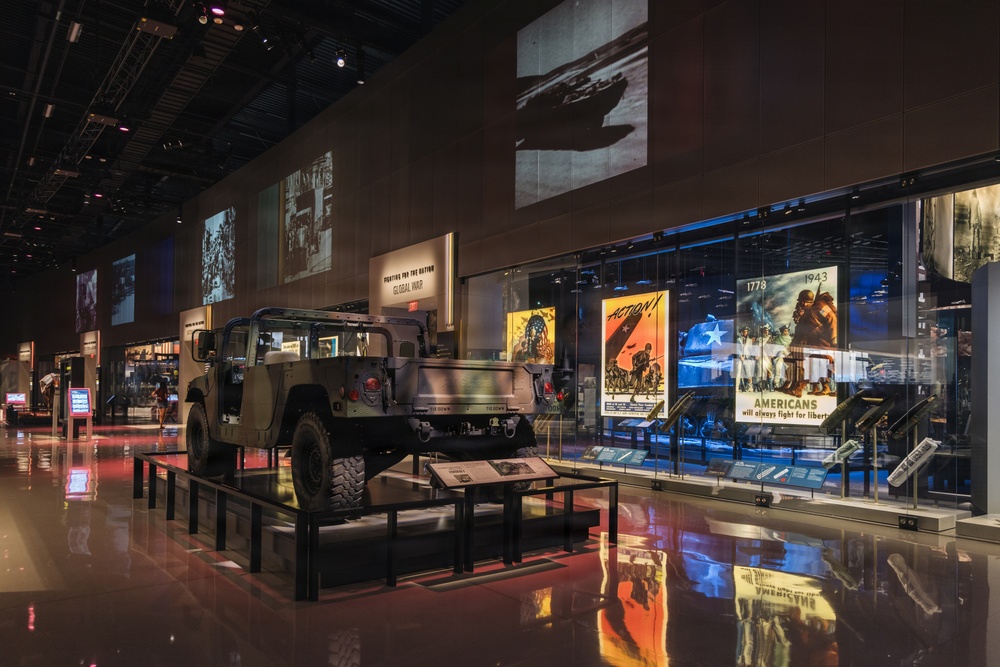A military museum exhibition hall featuring a prominently displayed HMMWV (Humvee) on a raised platform surrounded by historical military artifacts and propaganda posters. The exhibit spans multiple eras of American military history, with large projected images on the walls and illuminated display cases containing wartime posters and memorabilia. The space is dramatically lit with a mix of ambient and accent lighting on a reflective floor.