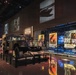 A military museum exhibition hall featuring a prominently displayed HMMWV (Humvee) on a raised platform surrounded by historical military artifacts and propaganda posters. The exhibit spans multiple eras of American military history, with large projected images on the walls and illuminated display cases containing wartime posters and memorabilia. The space is dramatically lit with a mix of ambient and accent lighting on a reflective floor.