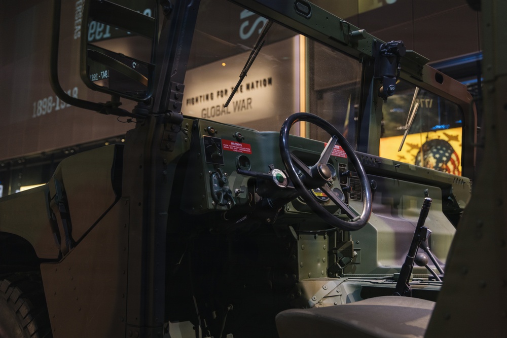 The image shows the interior cab of a military vehicle, likely a HMMWV (Humvee) or similar tactical truck, displayed in a museum setting. The vehicle is painted in olive drab green and features an open-door configuration with a visible steering wheel and dashboard controls. The museum backdrop includes signage reading 'FIGHTING FOR THE NATION' and 'GLOBAL WAR,' suggesting a military history exhibition.