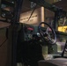The image shows the interior cab of a military vehicle, likely a HMMWV (Humvee) or similar tactical truck, displayed in a museum setting. The vehicle is painted in olive drab green and features an open-door configuration with a visible steering wheel and dashboard controls. The museum backdrop includes signage reading 'FIGHTING FOR THE NATION' and 'GLOBAL WAR,' suggesting a military history exhibition.