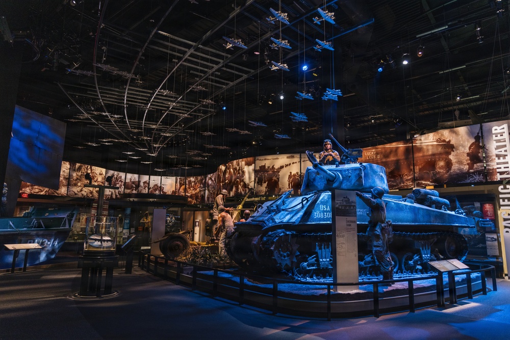 A dramatic museum exhibit hall, likely the National World War II Museum, featuring a prominently displayed M4 Sherman tank illuminated in blue lighting with the marking 'USA 3083083'. The exhibit includes suspended scale model aircraft hanging from the ceiling, large panoramic photographic murals on curved walls depicting WWII scenes, and life-size soldier figures positioned around the tank. The space is designed as an immersive theatrical environment combining artifacts, dioramas, and archival imagery.
