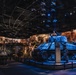 A dramatic museum exhibit hall, likely the National World War II Museum, featuring a prominently displayed M4 Sherman tank illuminated in blue lighting with the marking 'USA 3083083'. The exhibit includes suspended scale model aircraft hanging from the ceiling, large panoramic photographic murals on curved walls depicting WWII scenes, and life-size soldier figures positioned around the tank. The space is designed as an immersive theatrical environment combining artifacts, dioramas, and archival imagery.