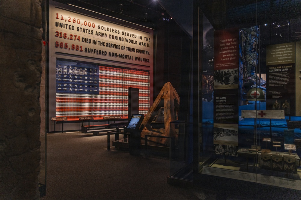 A museum exhibit dedicated to the United States Army's involvement in World War II. The centerpiece is a large illuminated display featuring an American flag made of soldier figurines, accompanied by statistics about soldiers who served, died, or were wounded. The exhibit includes artifacts, informational panels, and display cases related to the Army in Europe.