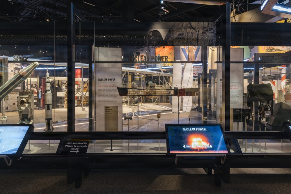 This image shows a museum exhibit dedicated to 'Nuclear Power,' featuring artifacts displayed behind glass panels including what appears to be a historic aircraft or seaplane and rocket/missile models. Interactive digital touchscreen displays are positioned in the foreground, inviting visitors to engage with the content. The exhibit is part of a larger museum floor that also references 'Space' in adjacent sections.
