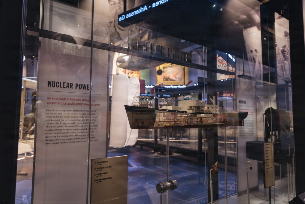 A museum exhibit dedicated to Nuclear Power, featuring a scale model of what appears to be the Army's MH-1A Sturgis floating nuclear power plant ship displayed in a glass case. The exhibit includes informational panels describing the Army Corps of Engineers' role in managing the world's first successful atomic energy program and the Manhattan Project. The display is part of a larger military history museum with additional exhibits visible in the background.