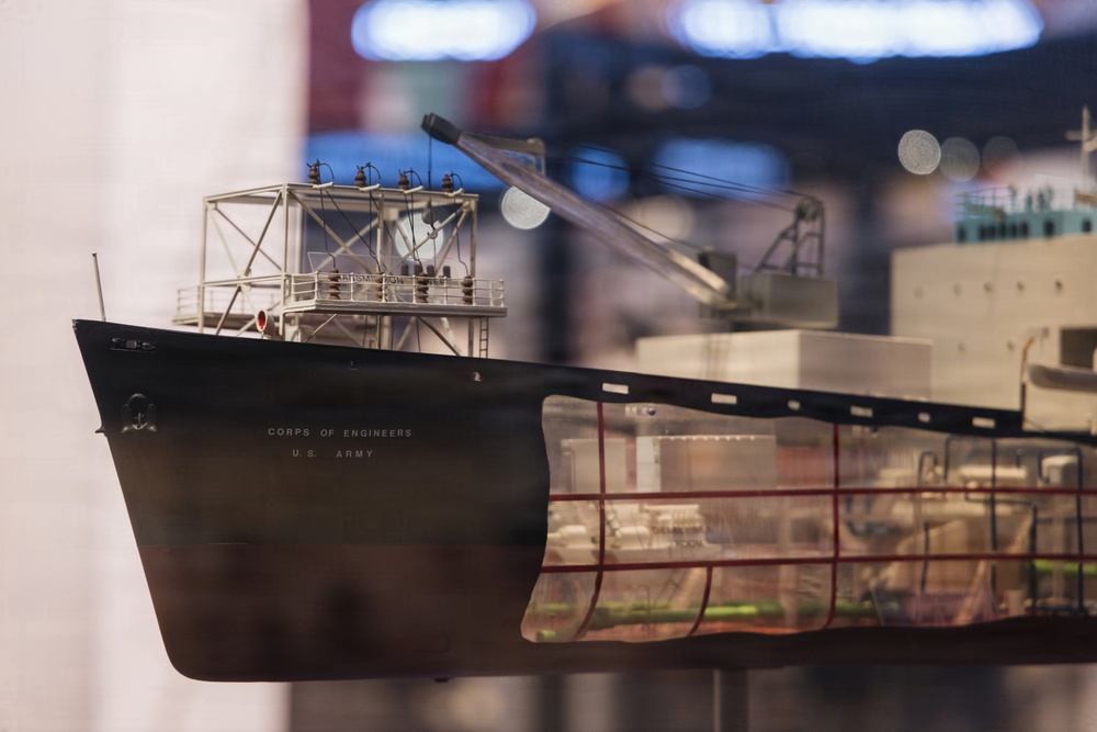 The image shows a detailed scale model of a U.S. Army Corps of Engineers vessel displayed in what appears to be a museum or exhibition setting. The model features a dark hull with white superstructure, crane equipment, and intricate deck details. A glass display case reflection reveals additional ship models in the background.